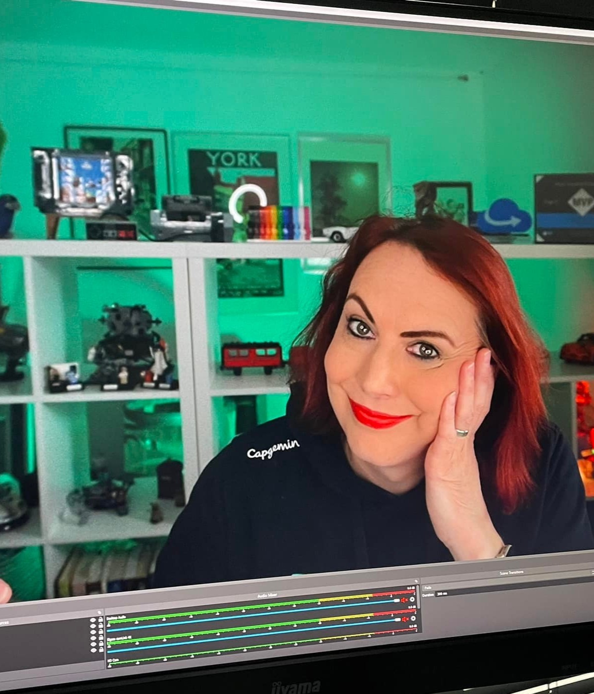 Stacy looking at the camera, she has red hair, and is resting on one arm. The outline of an OBS recording window can be seen so so we know this is being recorded. Behind her, in a mint green glow, are Lego sets on a white shelf
